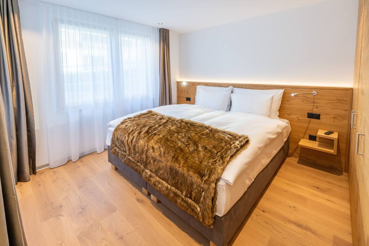 A modern hotel room with a wooden bed, a fur throw, a nightstand with a lamp, and a view of the city through sheer curtains.