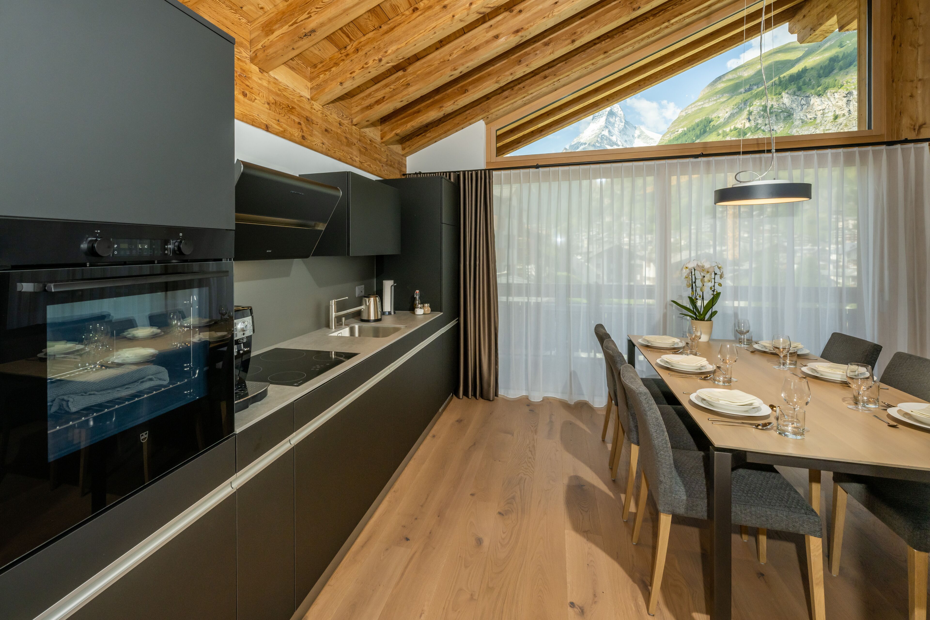 A modern kitchen with a built-in oven, a dining area with a table set for four, and a large window with a mountain view.