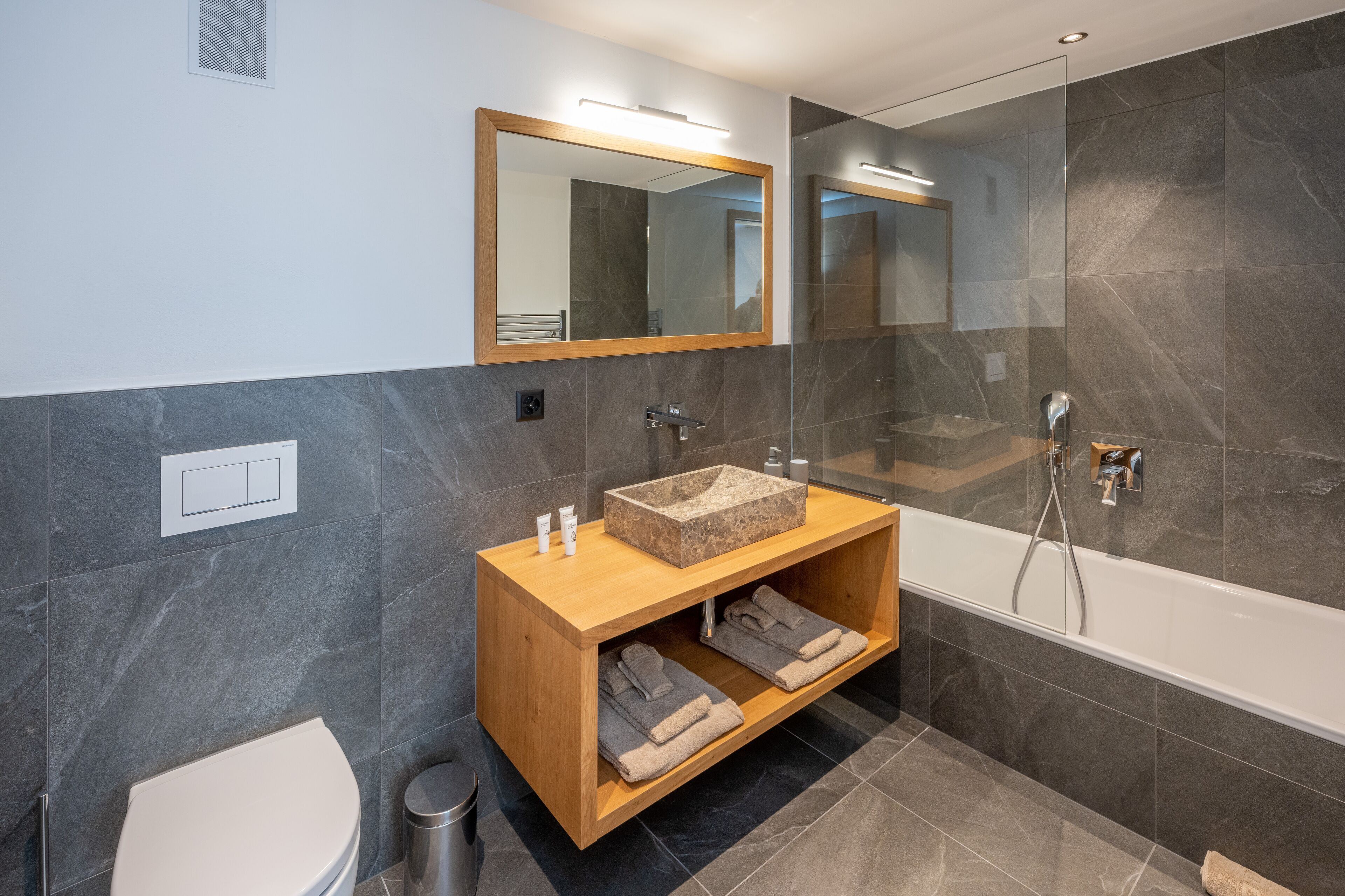 Ein modernes Badezimmer mit einem Holzwaschtisch, einem großen Spiegel, einer Badewanne und dunkel marmorierten Wänden.