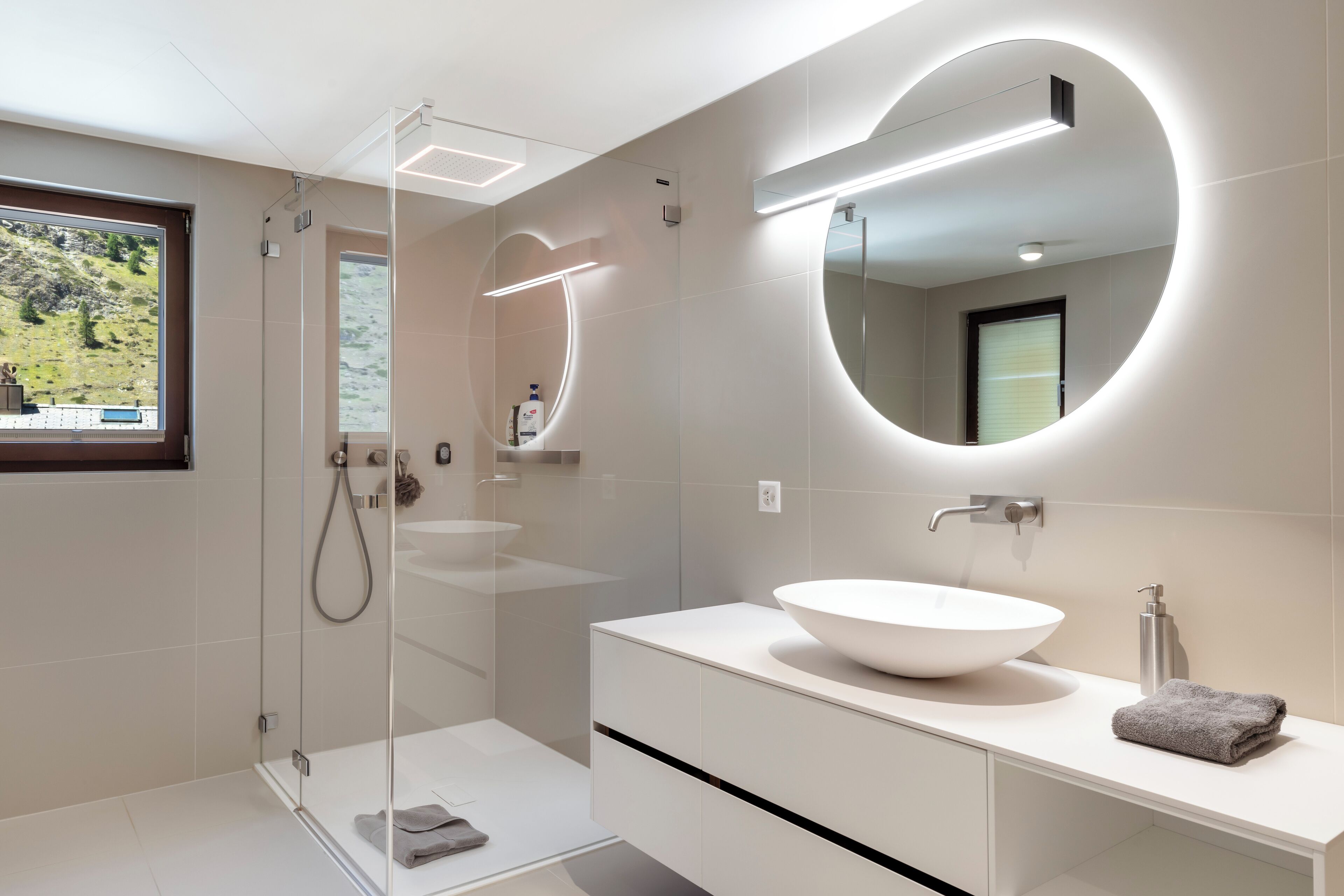 Ein modernes Badezimmer mit einer gläsernen Dusche, einem großen, ovalen Spiegel und einem weißen Waschbecken mit Aufsatzbecken.