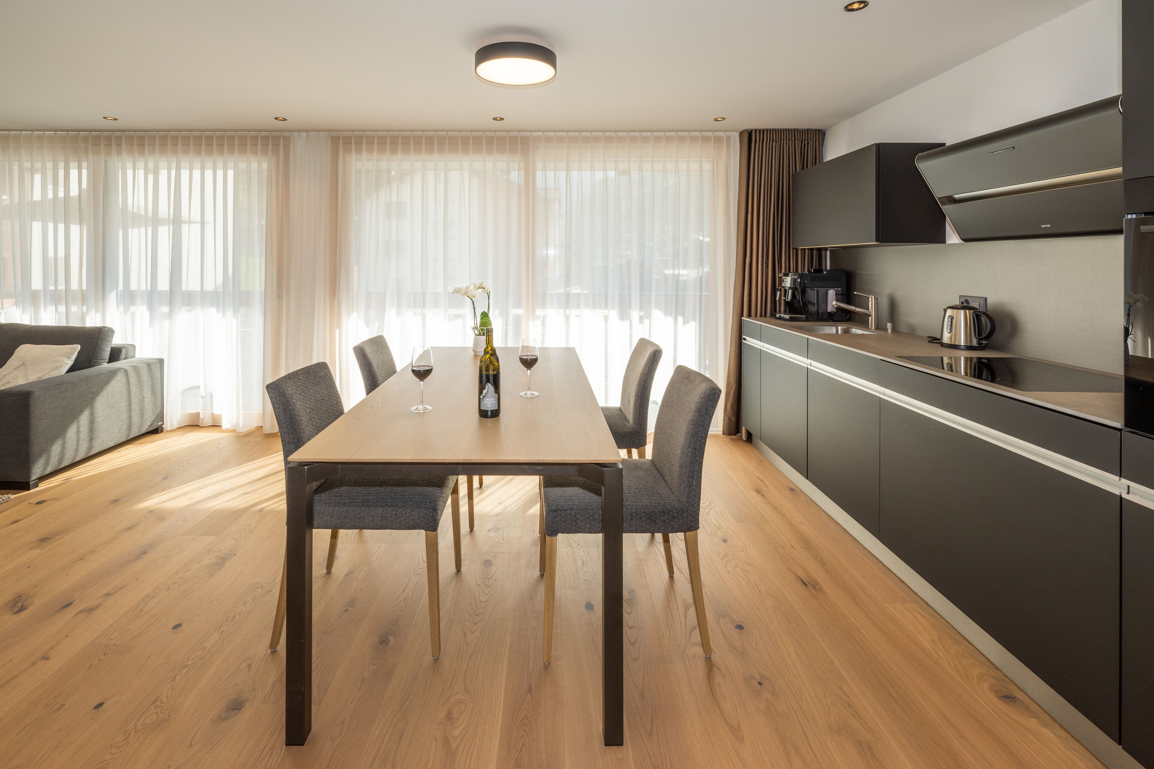 A modern dining area with a wooden table and chairs, a vase with flowers, and a kitchen with dark cabinets and a coffee machine.