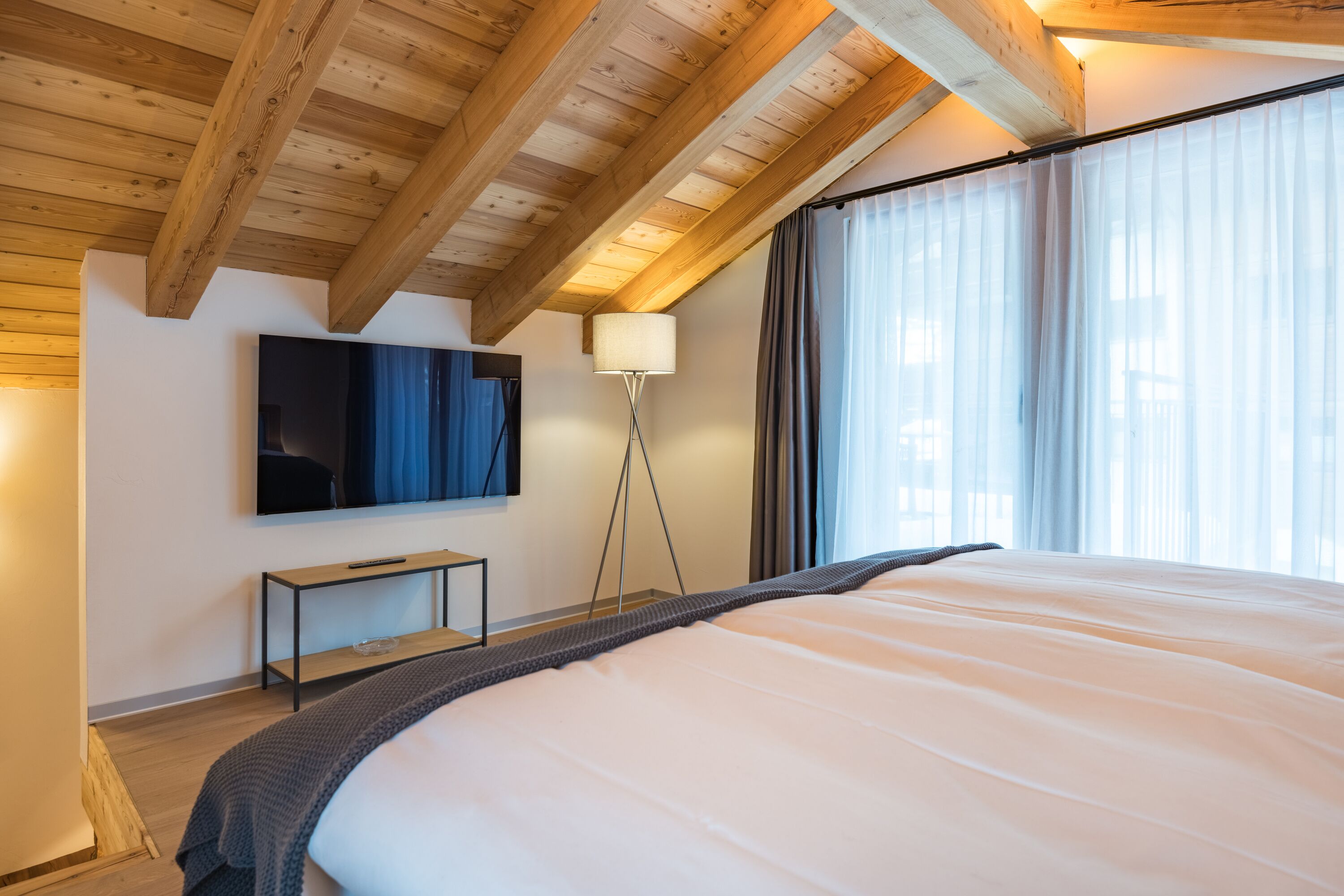 Een slaapkamer met een groot bed, een aan de muur bevestigde televisie, een nachtkastje en een vloerlamp.