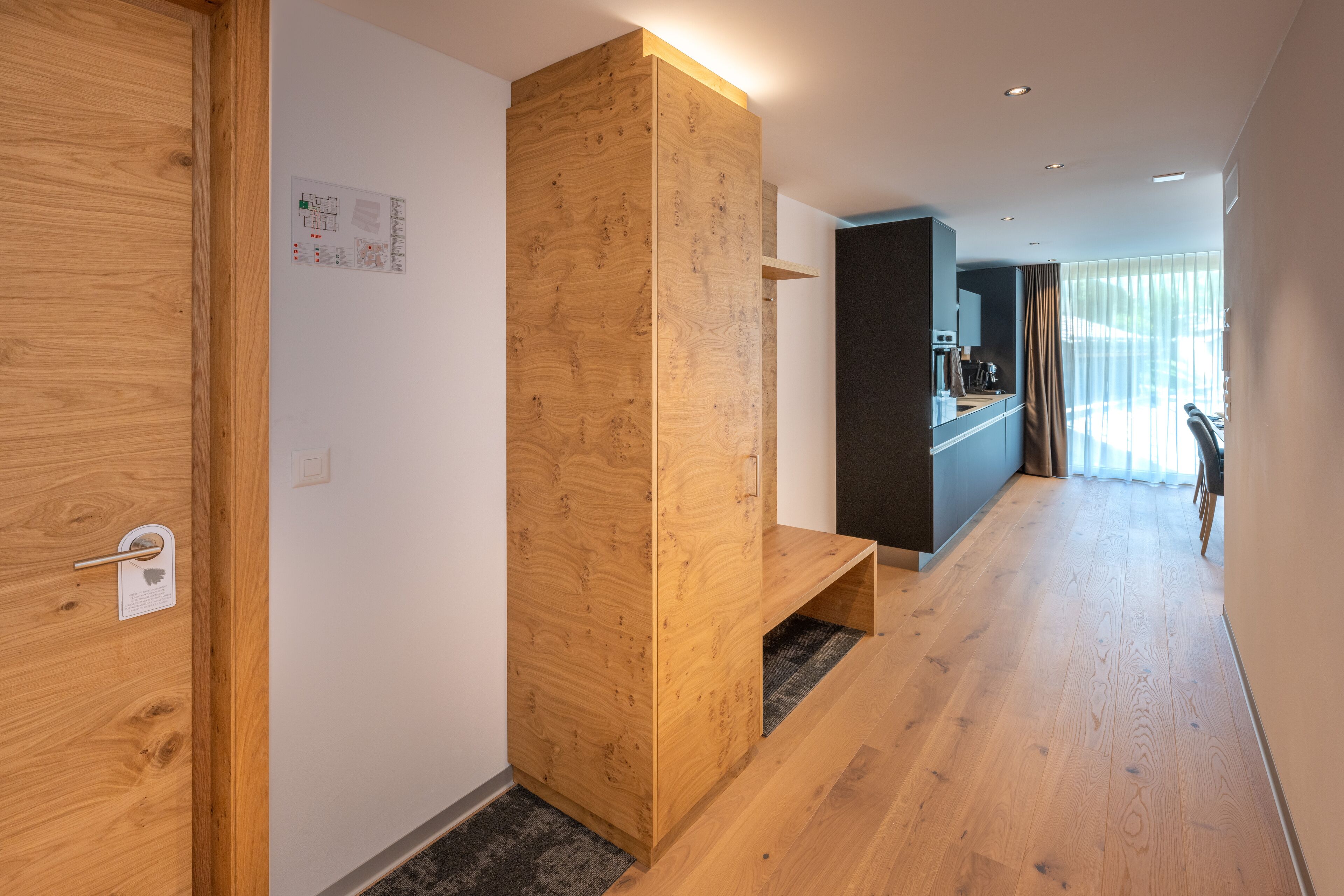 A modern hotel room with a wooden floor, a built-in bench, a desk, and a large window with curtains.
