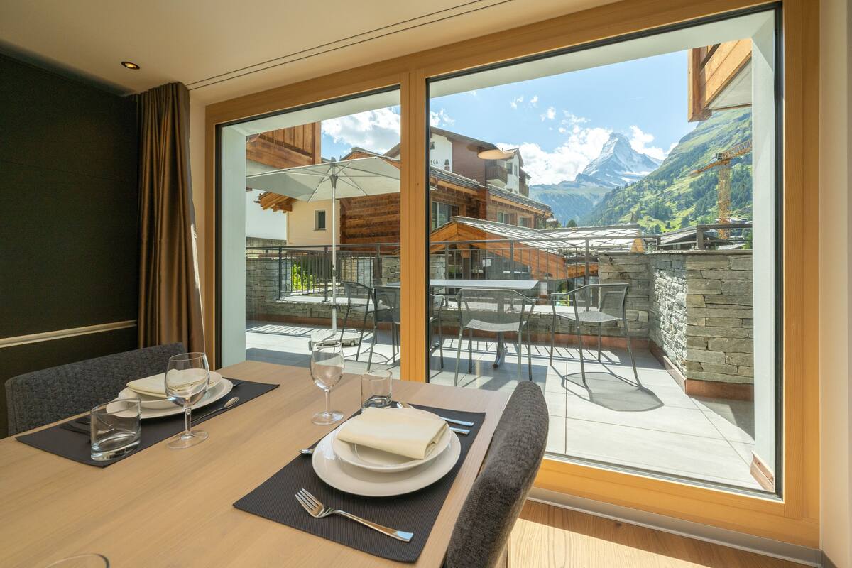 A dining area with a table set for two, a view of a mountain landscape through large windows, and outdoor seating with umbrellas.