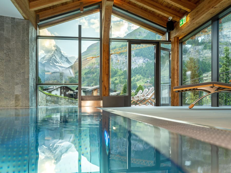 Una piscina interna moderna con vista diretta sulle montagne e su un cantiere edile attraverso ampie finestre.