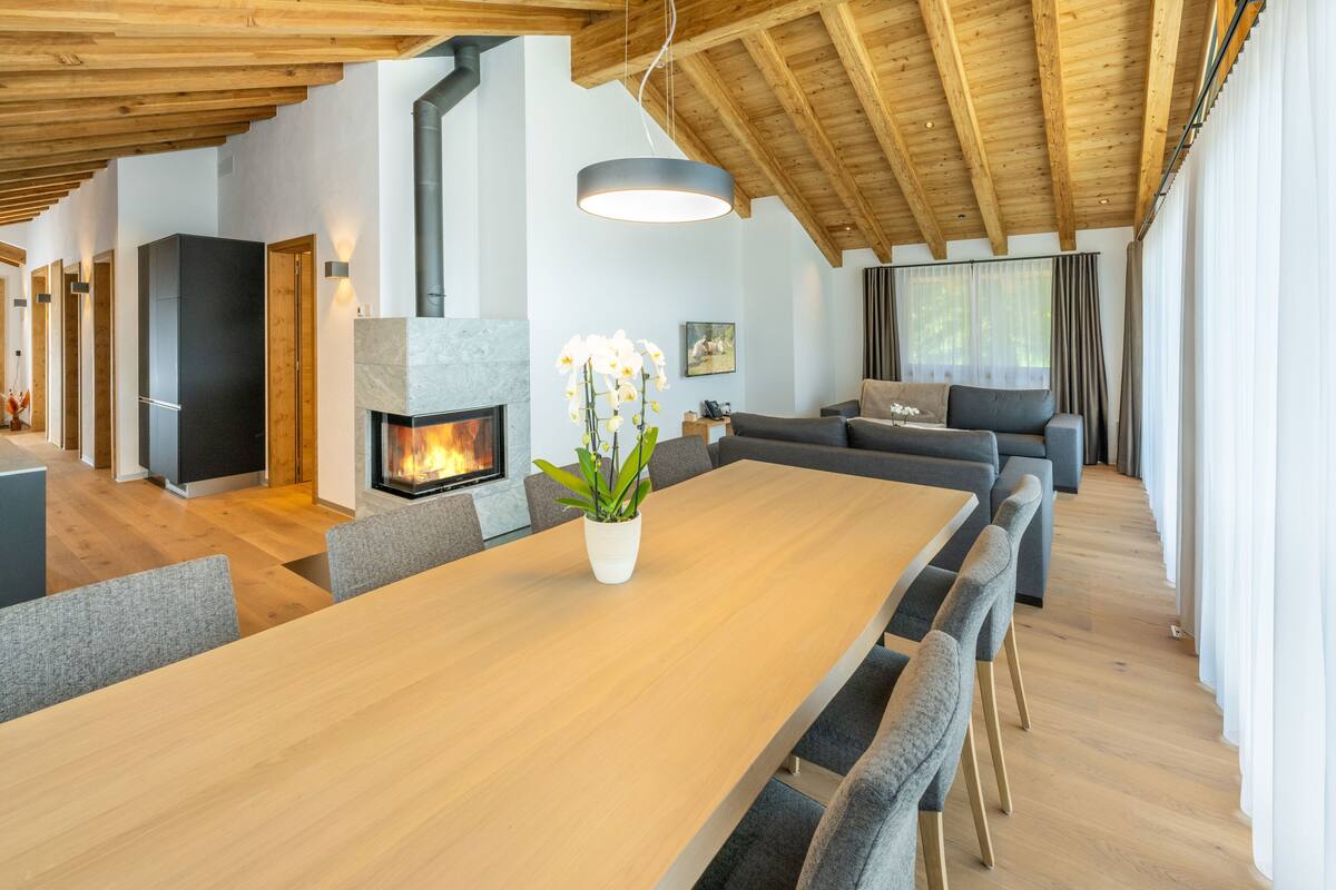 A spacious living area with a wooden ceiling, a fireplace, a dining table, and grey upholstered seating.