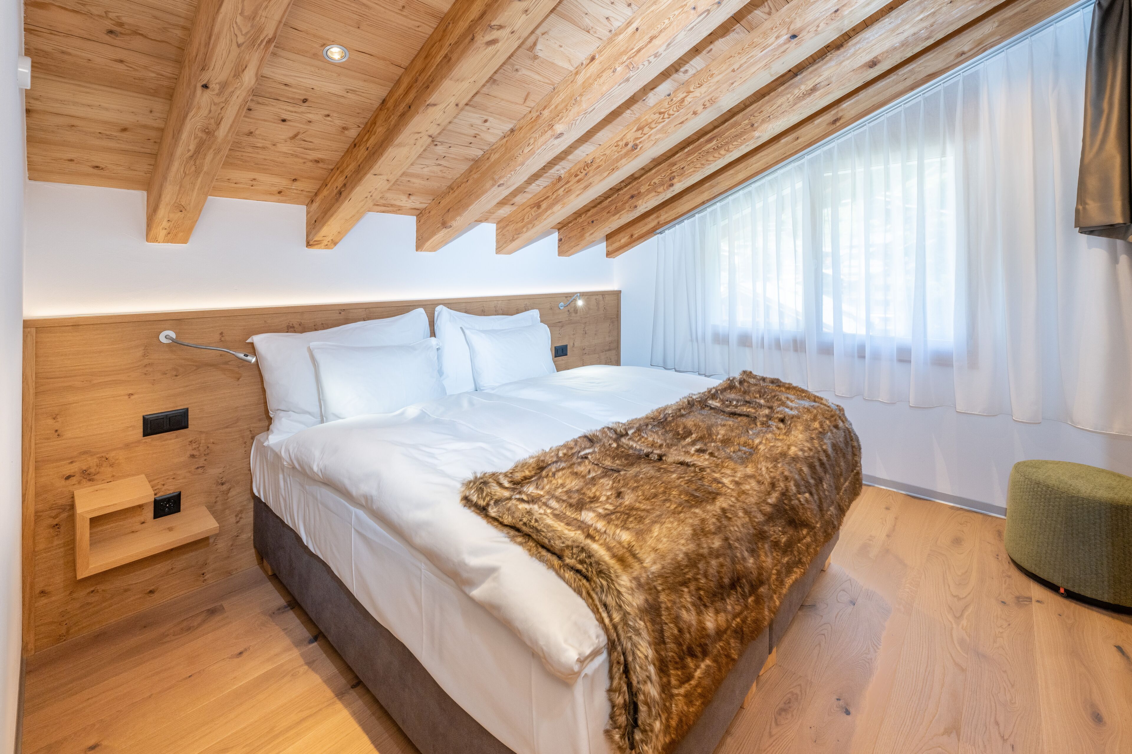 Ein modernes Schlafzimmer mit Holzboden, einem großen Bett mit weißer Bettwäsche, einem Fellüberwurf und einem grünen Hocker.