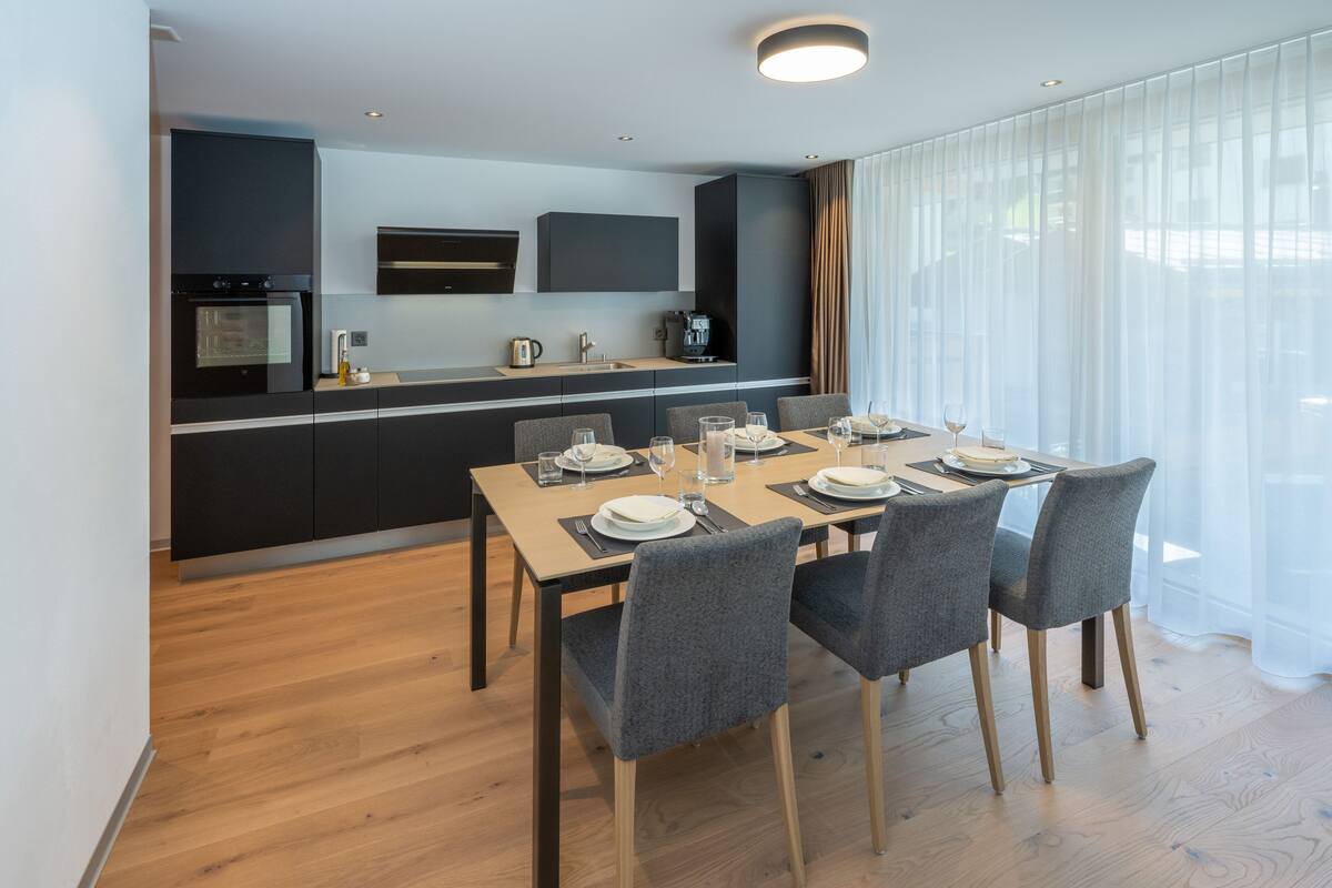 A modern dining area with a wooden table set for four, a kitchen in the background, and large windows with sheer curtains.