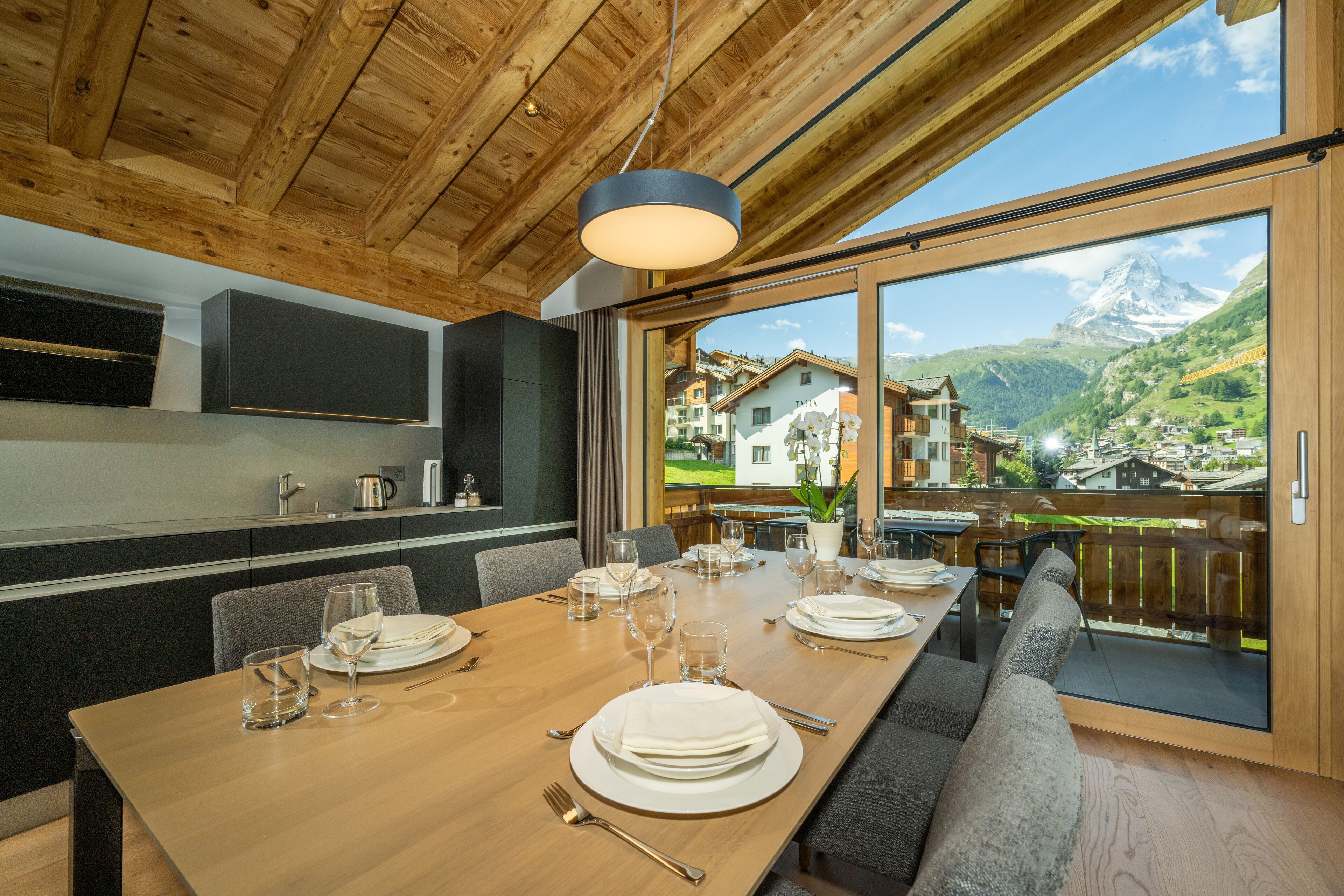 A dining area with a wooden table set for a meal, a large window with a mountain view, and a modern kitchen with built-in appliances.