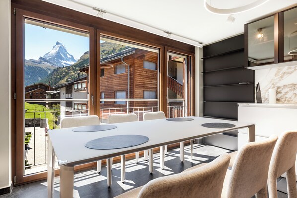 Quarto de hotel moderno com uma grande mesa de jantar, assentos confortáveis, vista para uma montanha e uma varanda com fachada de madeira.