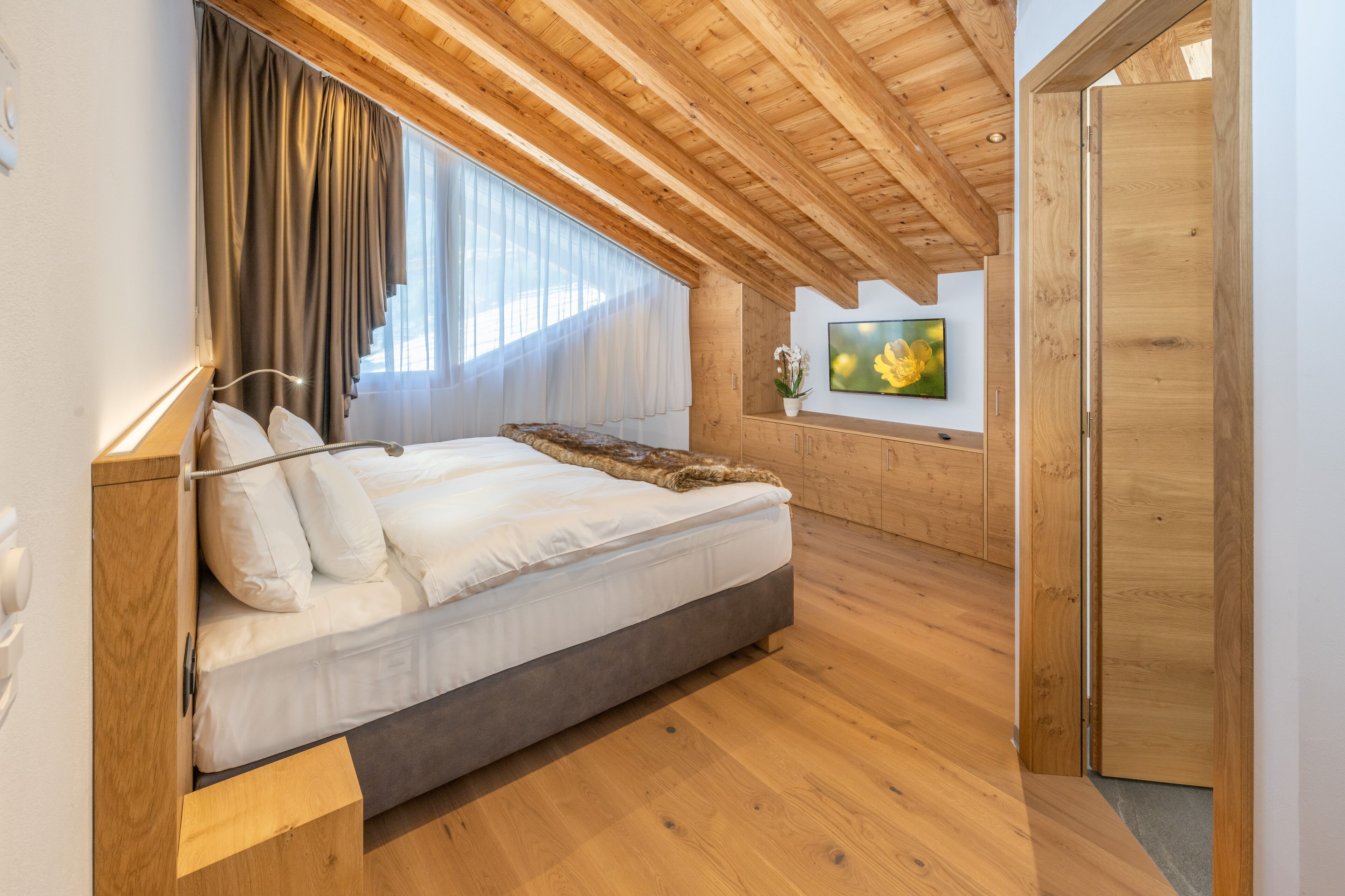 A modern bedroom with a large bed, wooden ceiling beams, a wooden wardrobe, and a large window with curtains.