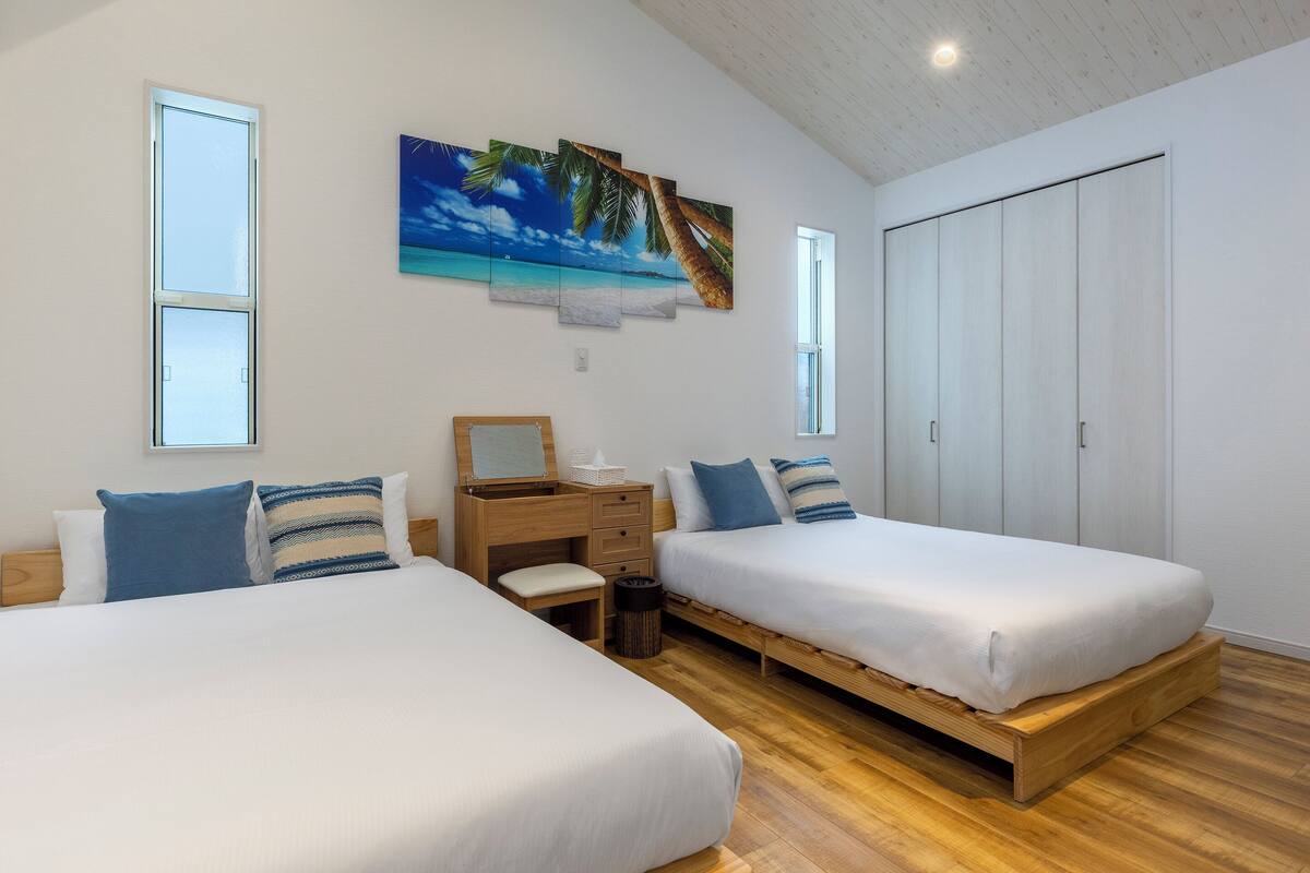 A bedroom with two beds, a desk, a chair, a trash bin, and a wardrobe.