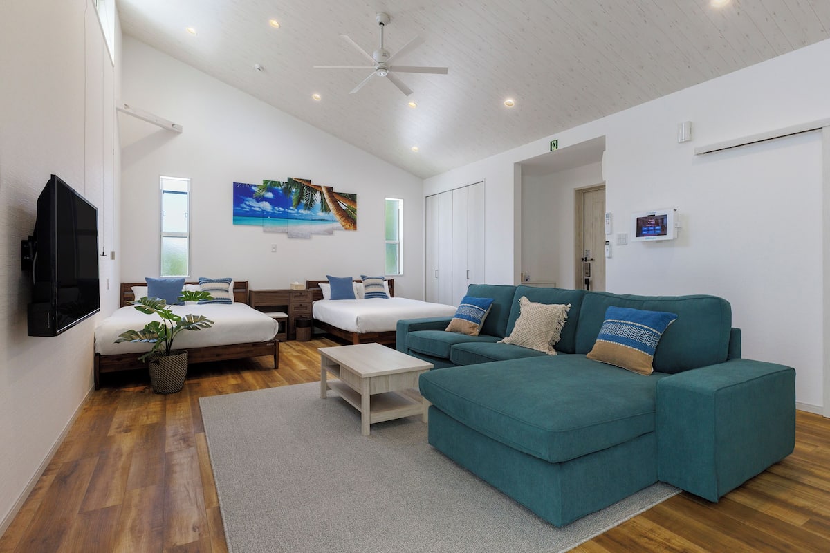A spacious living area with a teal sofa, a white coffee table, a TV mounted on the wall, and a ceiling fan.