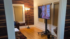 Una habitación con piso de madera, un televisor sobre un pedestal, un escritorio con una silla y una lámpara.