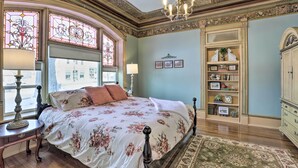 Ein Schlafzimmer mit floralem Bettbezug, einer Kristallleuchter, einem Bücherregal und einem großen Fenster mit Buntglas.