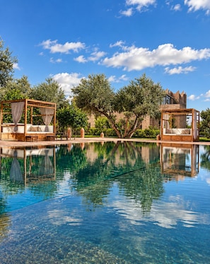 A clear pool with two wooden canopy beds, one on each side, and a castle-like building in the background.
