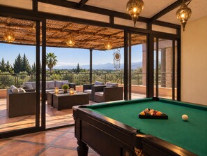 A pool table with a green felt surface, surrounded by wicker furniture and a view of a mountainous landscape through large windows.