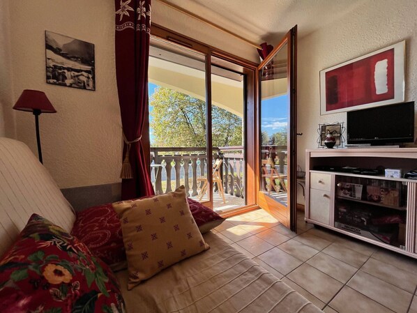 A cozy room with a sofa, a television, and a balcony with a view of trees.