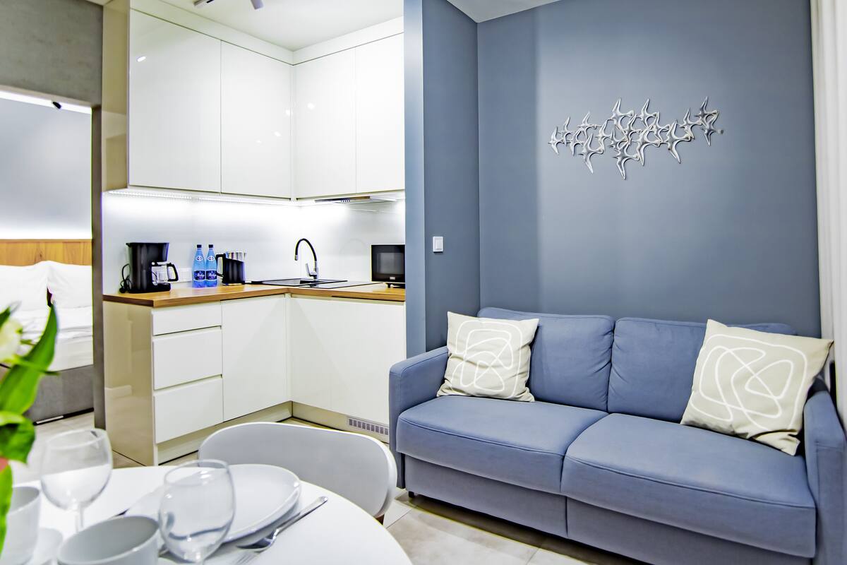 Een moderne woonkamer met een blauwe bank, een eettafel gedekt met borden en glazen, en een keuken met witte kasten en een koffiezetapparaat.