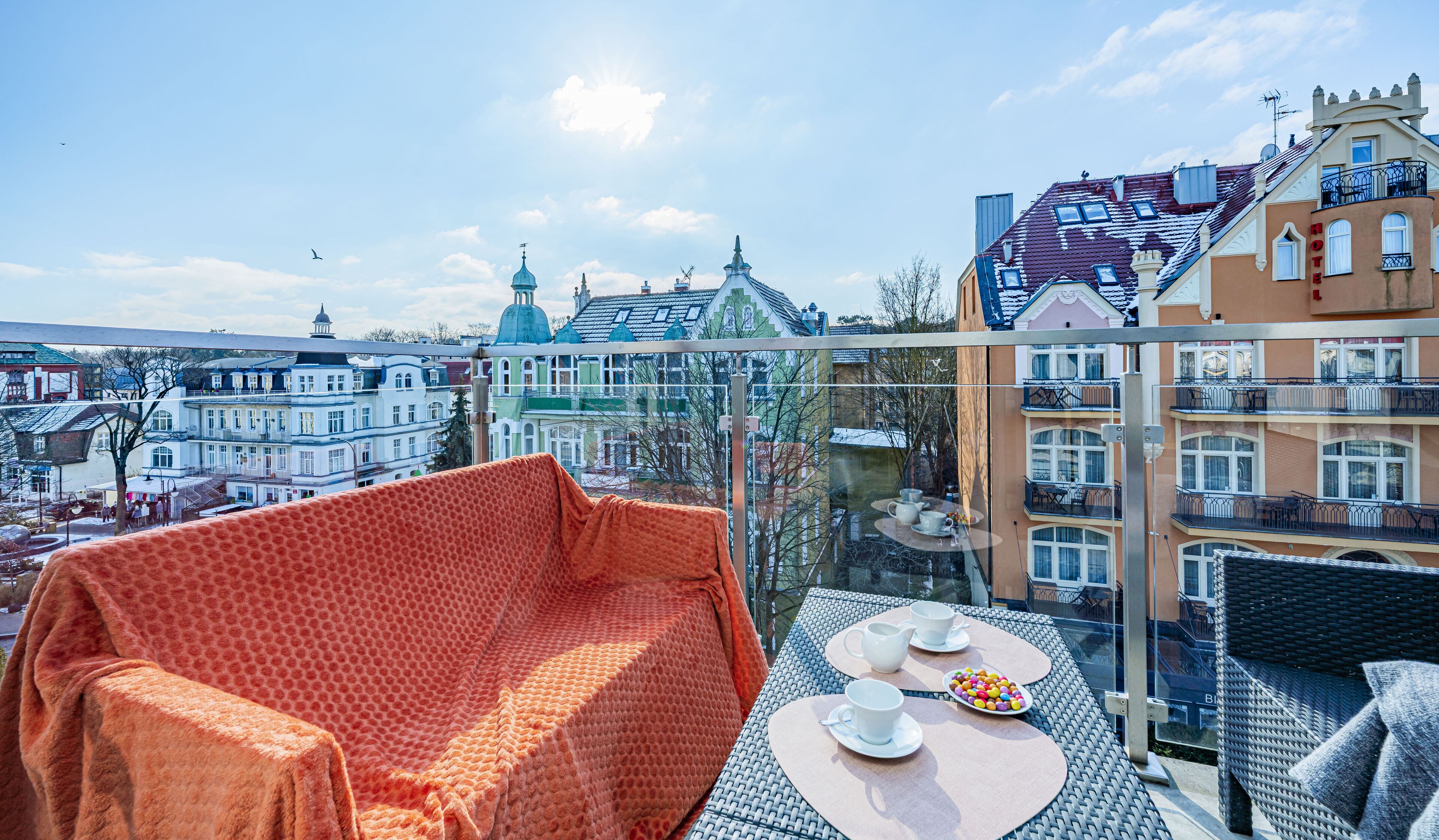 Een balkon met een rood gestreept bankje, een ronde tafel met kopjes en een bord met snoepjes, en een glazen balustrade met uitzicht op een stadsbeeld met historische gebouwen.
