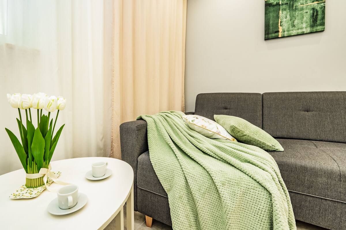 Een woonkamer met een grijze bank, een groene deken en een vaas witte tulpen op een witte tafel.