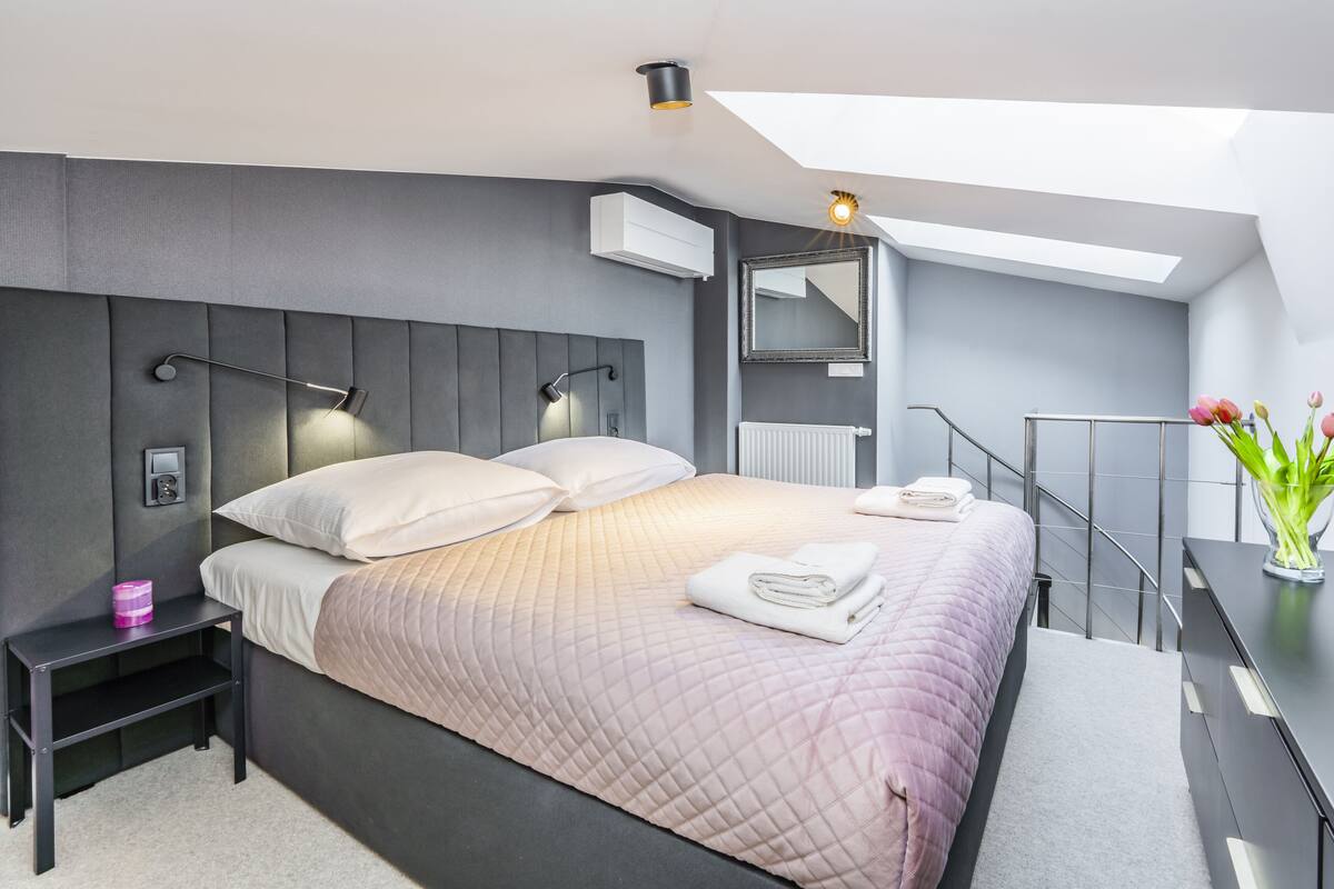 Een moderne slaapkamer met een groot bed, bedlampen, een nachtkastje, een spiegel, een radiator, een televisie en een vaas met bloemen.