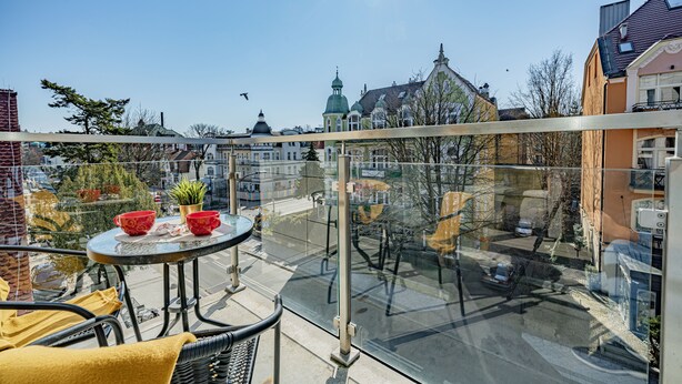 Un balcon avec une barrière en verre, une petite table ronde avec des tasses rouges et une plante en pot.