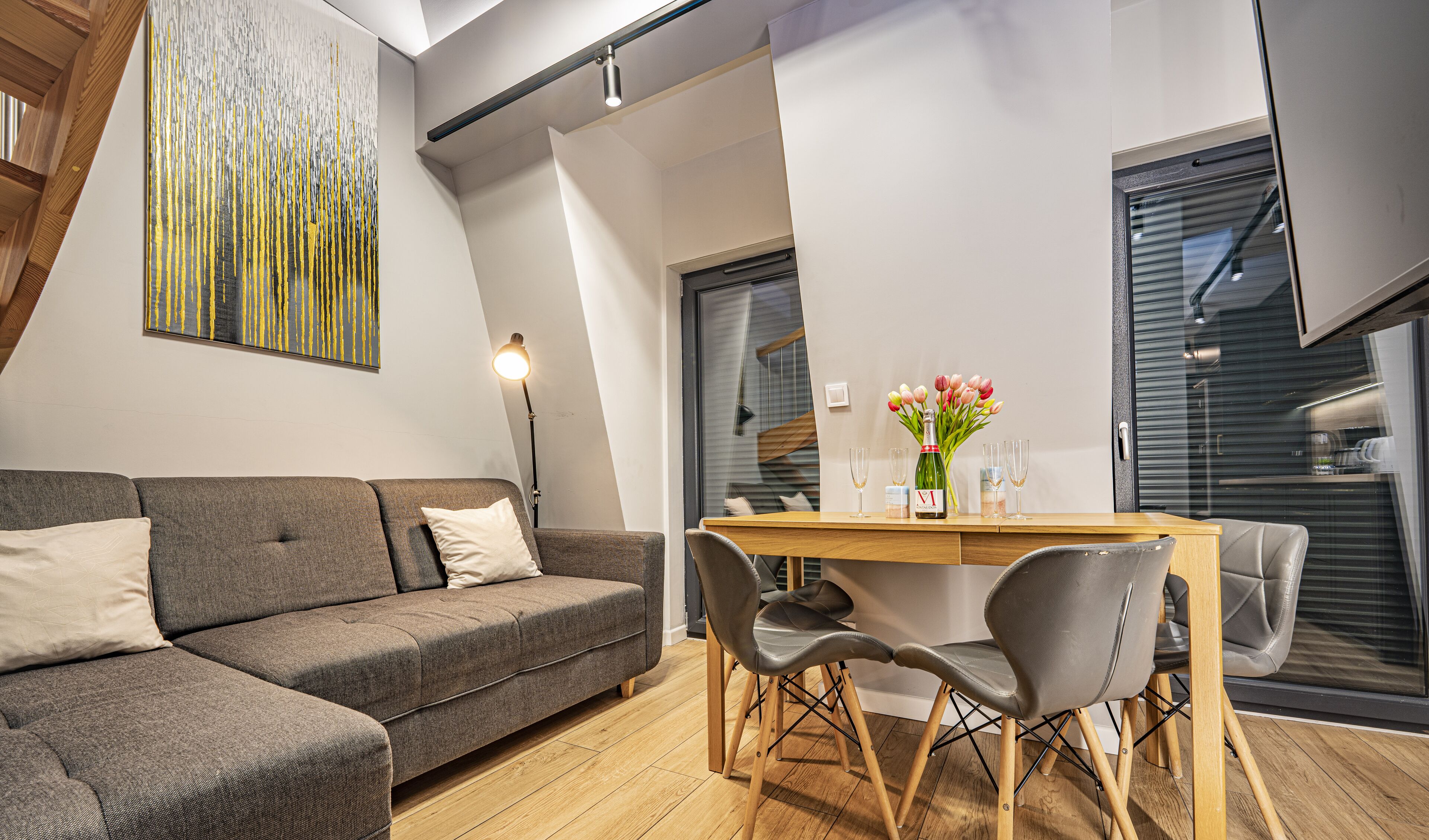 Ein modernes Wohnzimmer mit grauem Sofa, Holzesstisch und Stühlen. An der Wand hängt ein Gemälde und es gibt ein Fenster mit Jalousien.