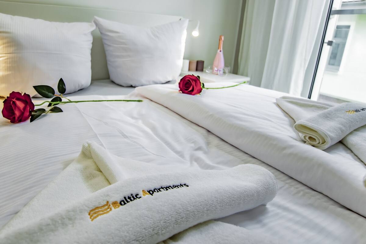 Een net opgemaakt bed met witte lakens, twee rode rozen en een opgerolde handdoek met de tekst 'Baltic Apartments' erin geborduurd.