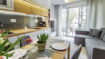 A modern living room with a grey sofa, a dining table set with plates and cutlery, and a kitchen area with wooden cabinets and a coffee maker.