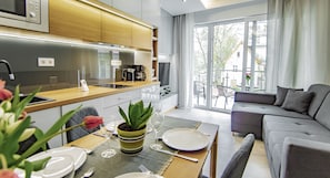 A modern living room with a grey sofa, a dining table set with plates and cutlery, and a kitchen area with wooden cabinets and a coffee maker.