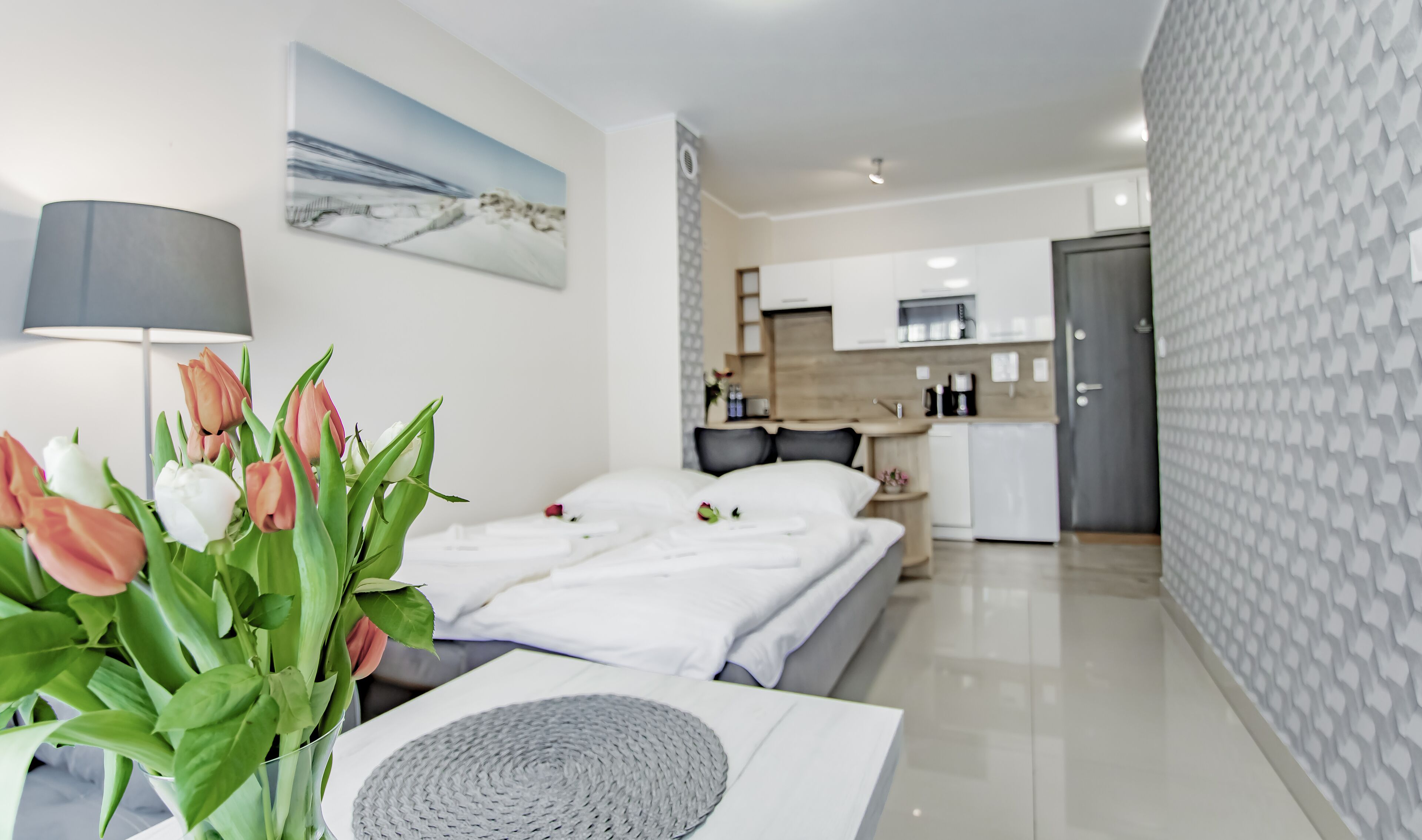 Una habitación de hotel moderna con una cama, una mesita de noche con lámpara y flores, una zona de cocina y una pared con diseño.