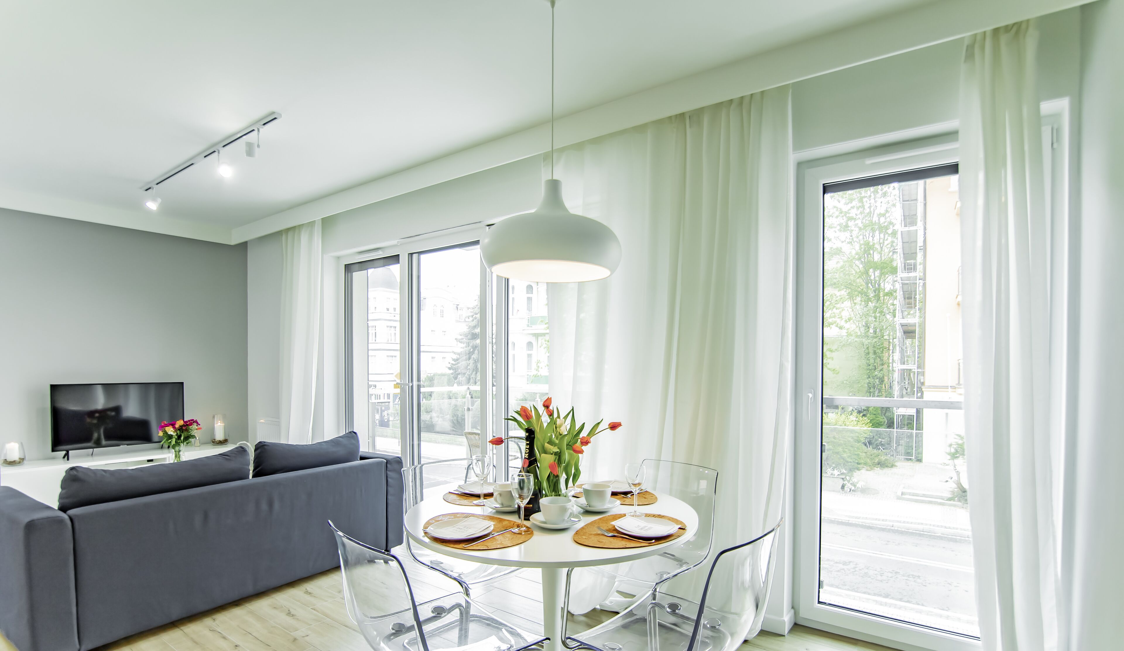 A modern living room with a grey sofa, a dining table set for four, and large windows with white curtains.