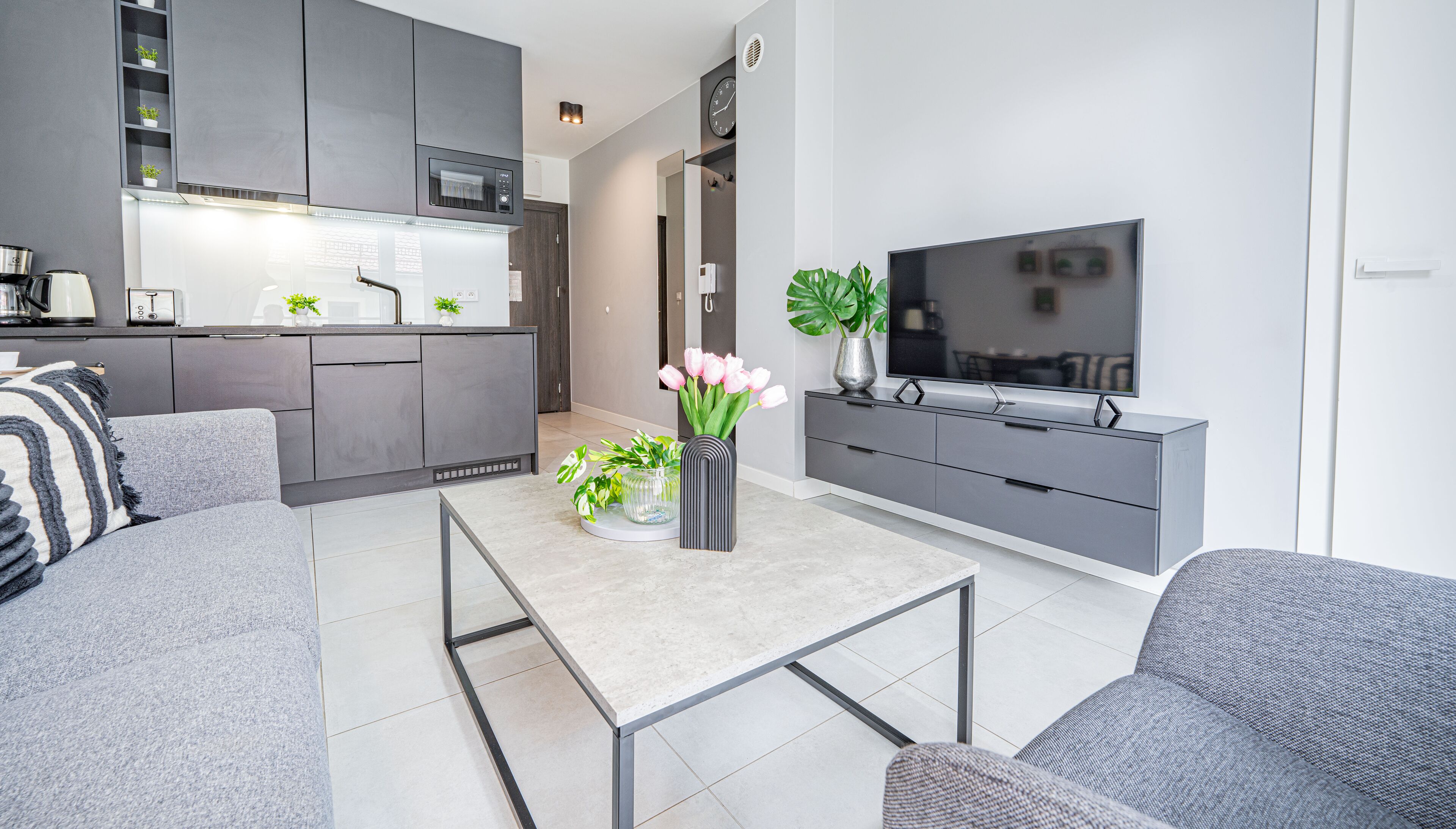 Un salon moderne avec un canapé gris, une table basse et un téléviseur posé sur un meuble gris. On aperçoit au fond une cuisine avec des meubles gris et un four encastré.