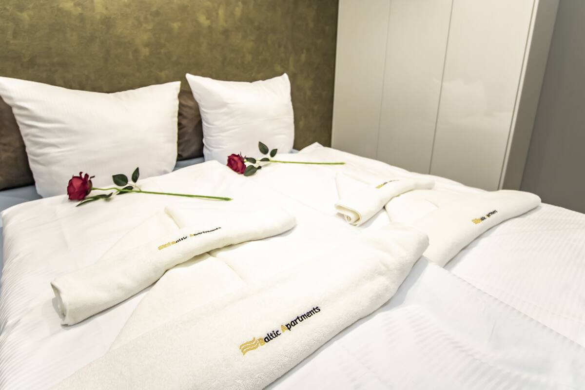 Een net opgemaakt bed met witte lakens, een roos en handdoeken met de branding 'Baltic Apartments'.