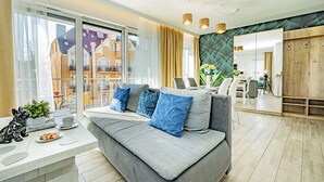 Un salon moderne avec un canapé gris, des coussins bleus, une petite table sur laquelle se trouvent une plante et une assiette de biscuits, et une vue sur le balcon extérieur.