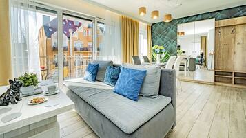 A modern living room with a grey sofa, blue cushions, a small table with a plant and a plate of cookies, and a view of the outside balcony.