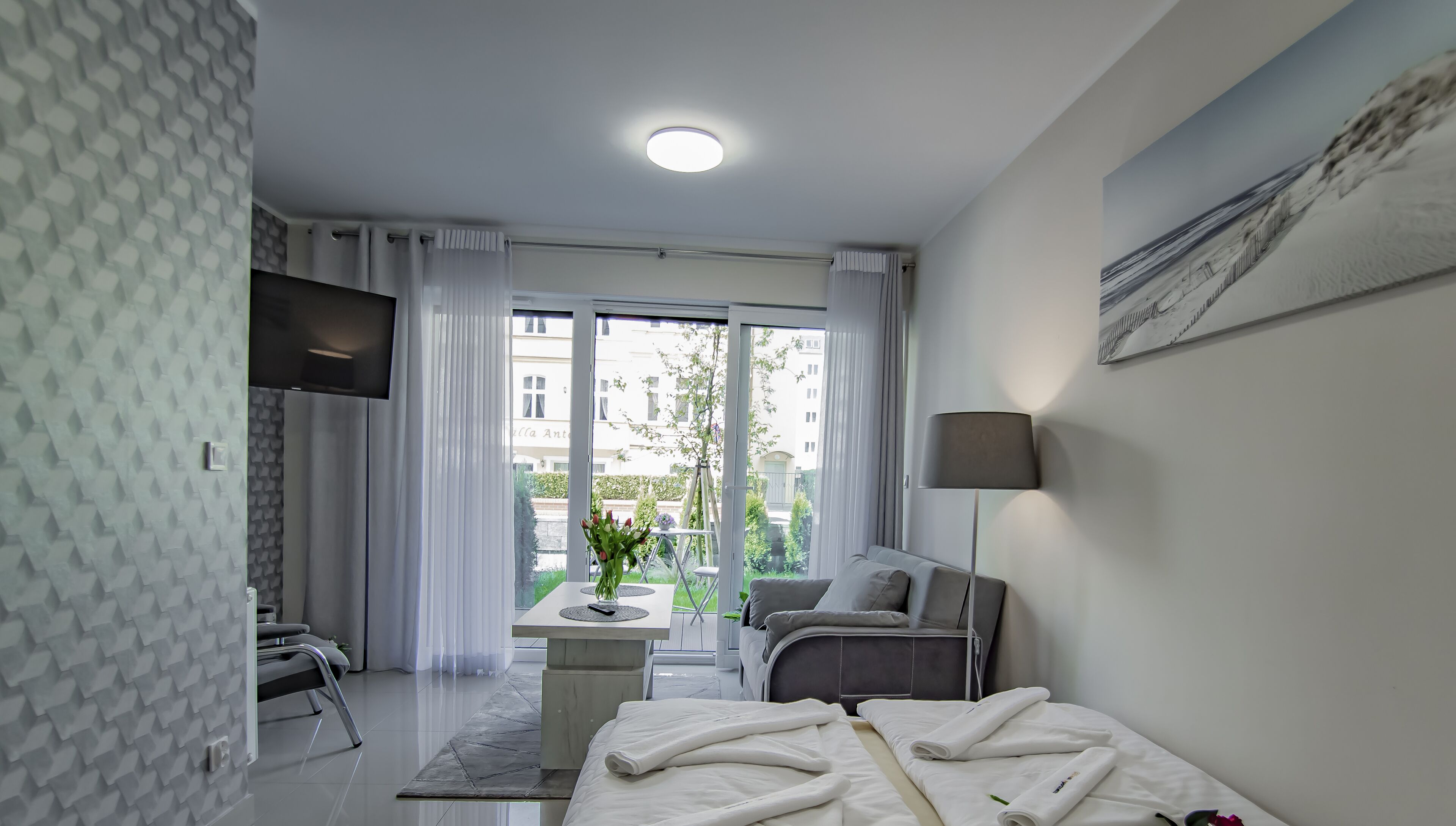 Quarto de hotel moderno com cama, escrivaninha, cadeira, televisão e varanda com vista para edifícios e vegetação.