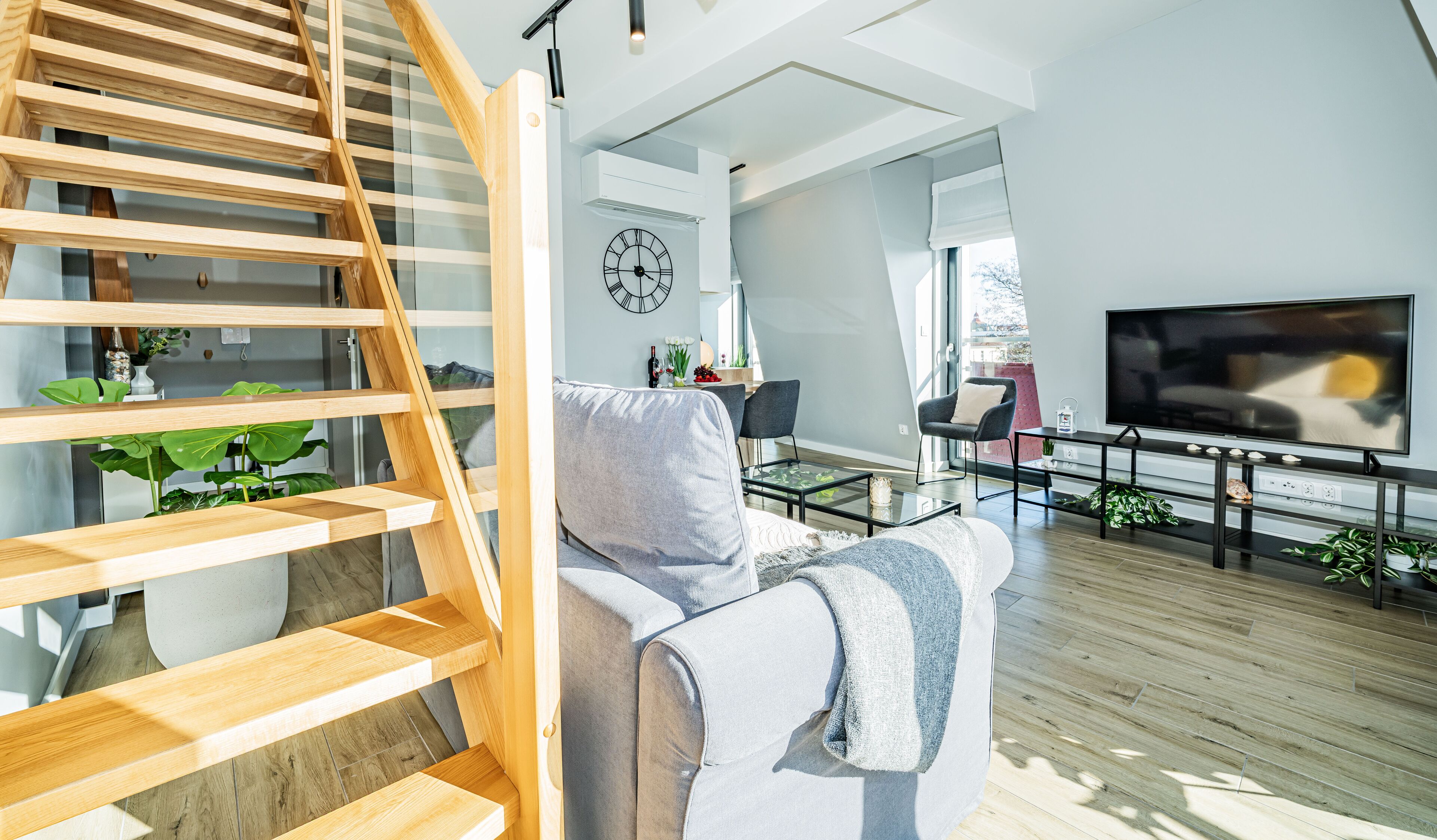 A modern living room with a wooden staircase, a grey sofa, a flat-screen TV, and a dining area with a table and chairs.