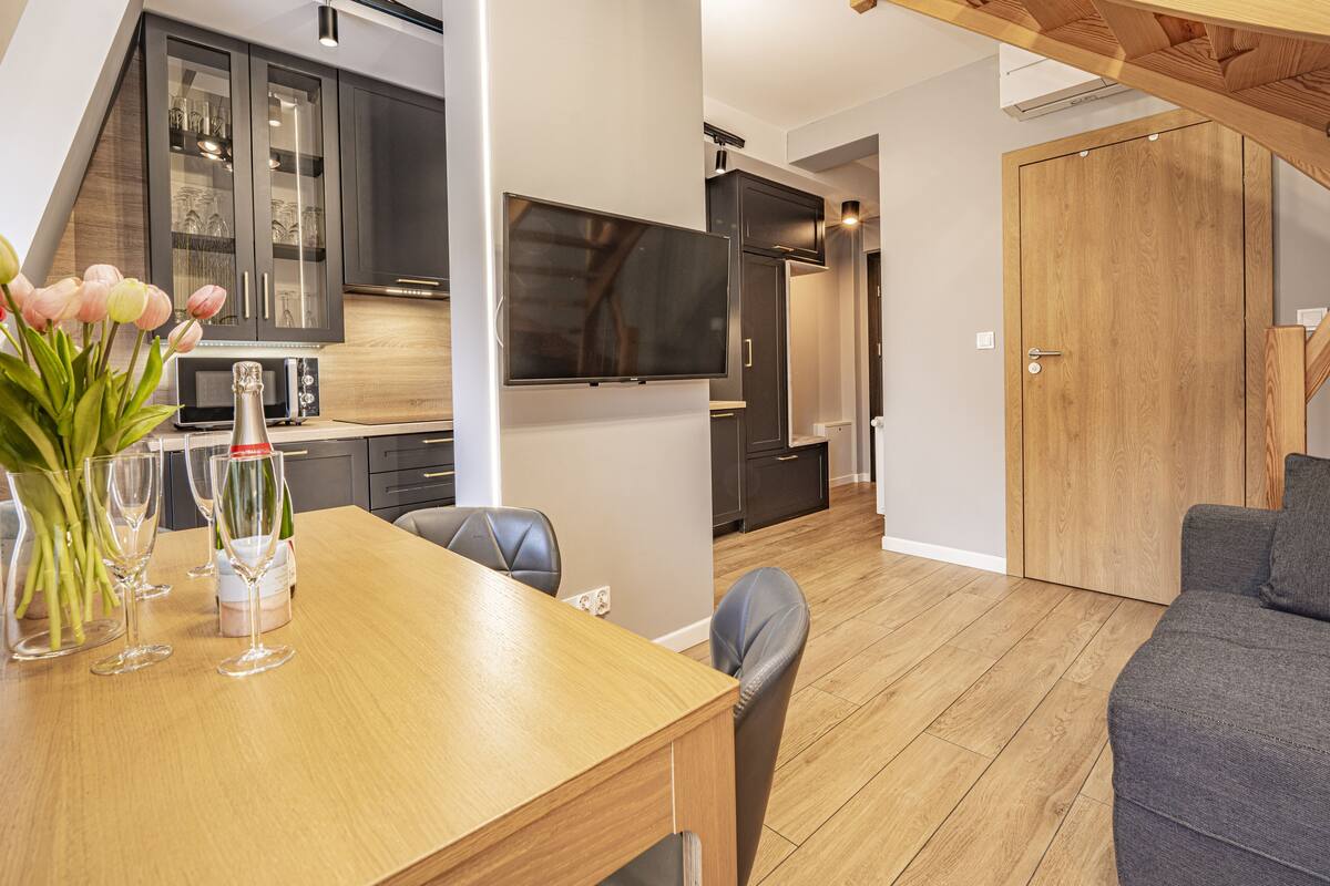 Een moderne keuken met donkere kasten, een inbouwoven en een flatscreen televisie. Er is een eettafel met stoelen en een bloemenvaas.
