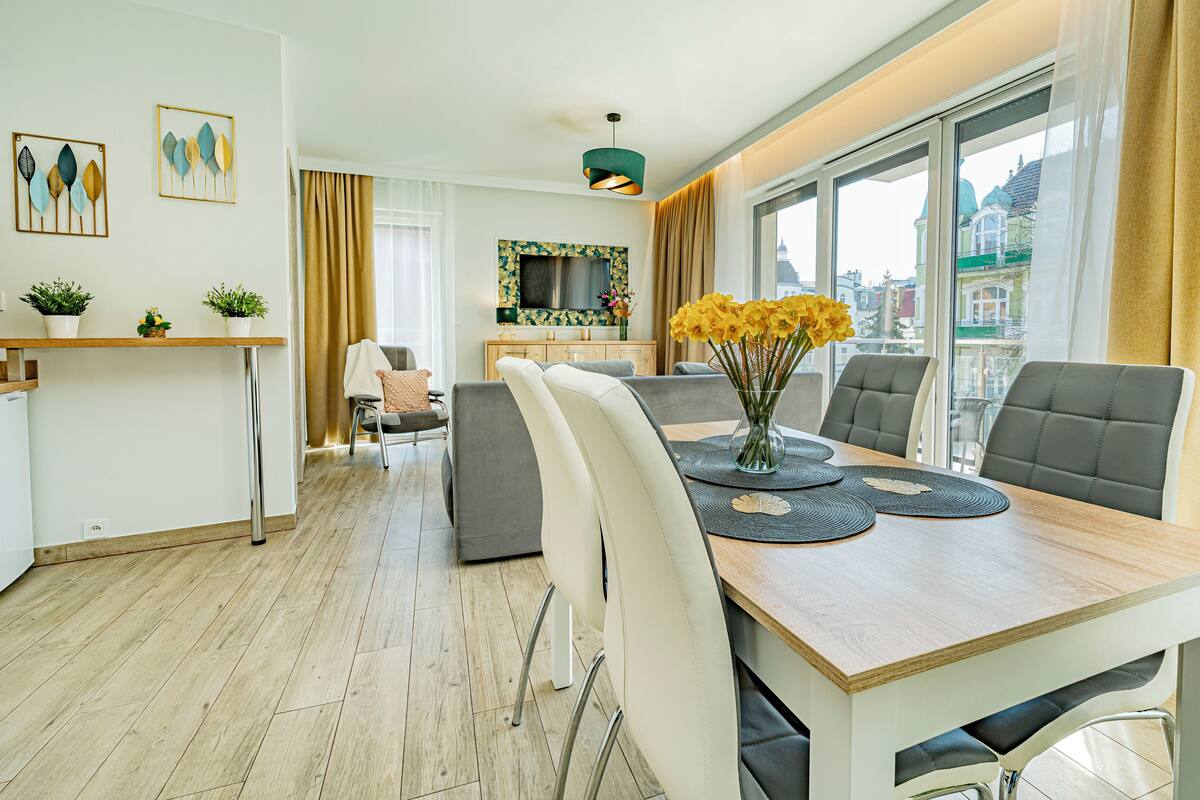Een moderne eetruimte met een houten tafel, grijs gestoffeerde stoelen en een vaas met gele bloemen.
