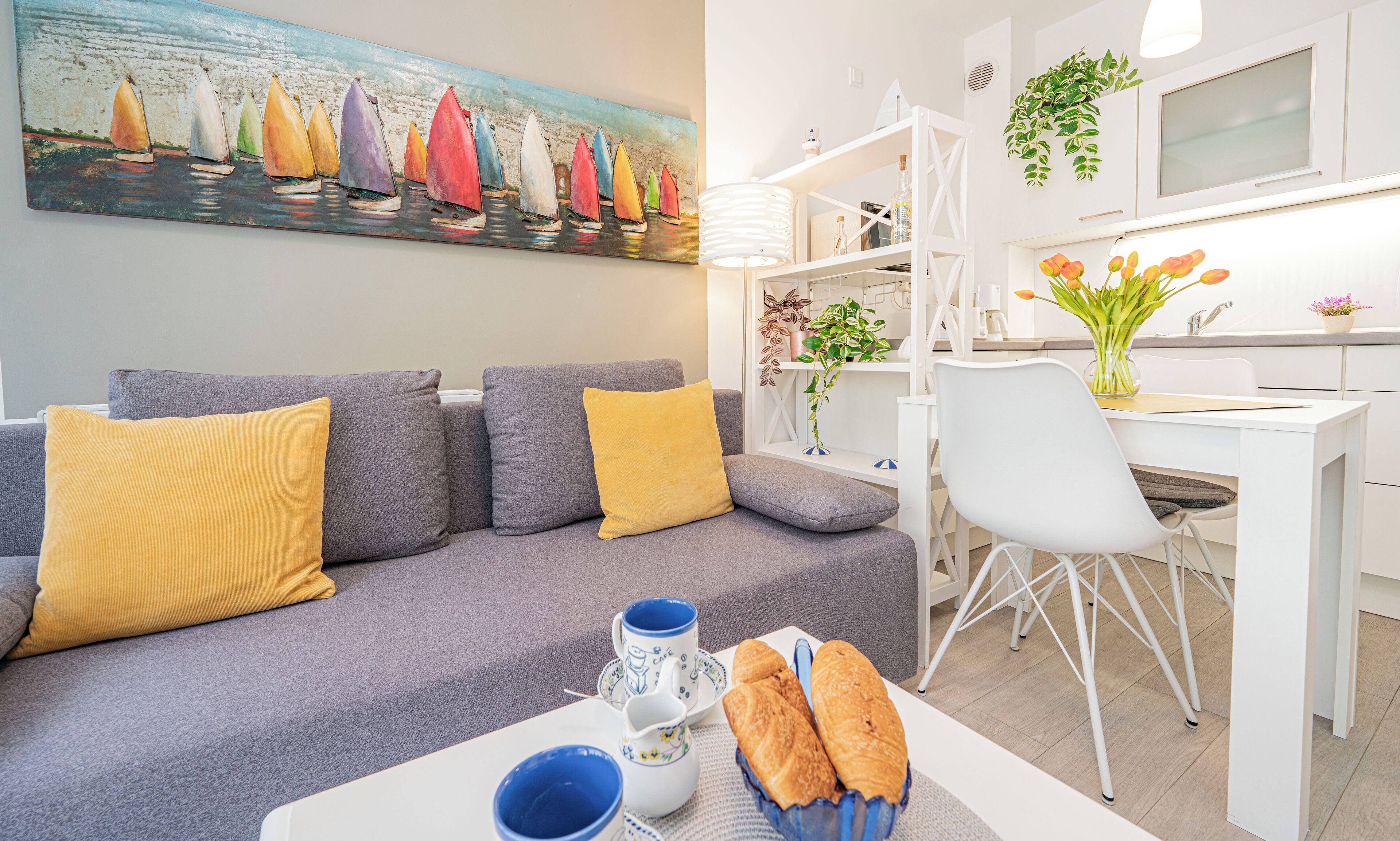 Een moderne woonkamer met een grijze bank, gele kussens, een eettafel met een witte stoel, en een schilderij van kleurrijke zeilboten aan de muur.