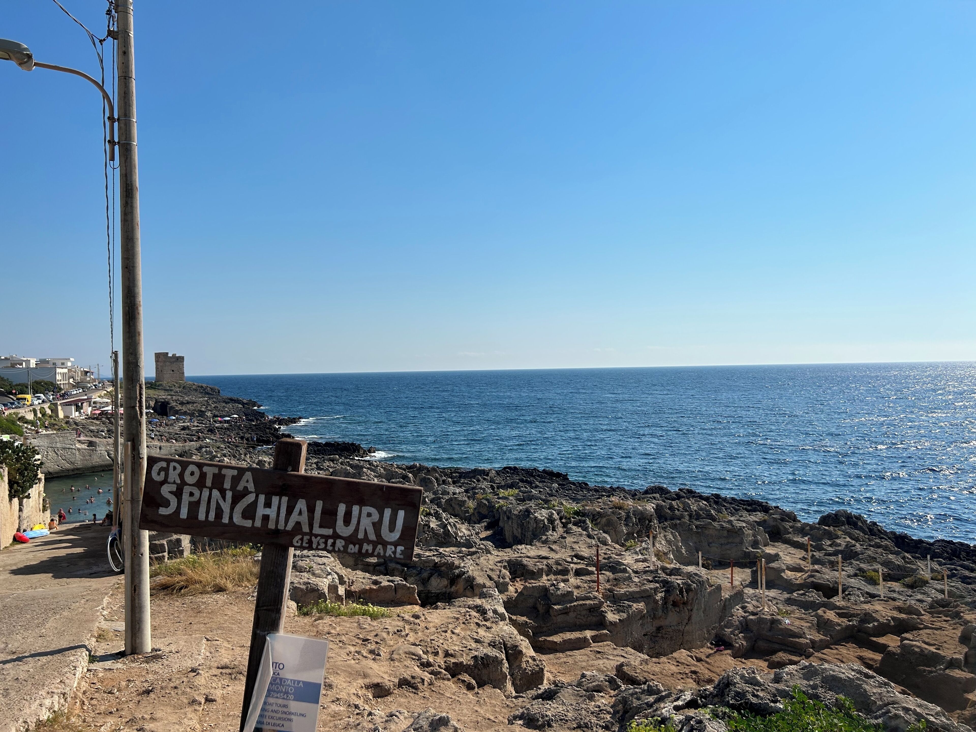 Un panneau indicateur portant les inscriptions « Grotta Spinichaluru » et « Geyser di Mare », surplombant une côte rocheuse avec la mer en arrière-plan.