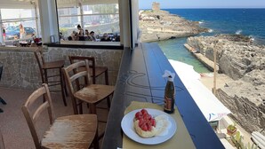 A terrace with stone walls, a table with a plate of food, and a view of the sea and rocky coastline. - Seafront historical villa with fabulous garden and private pool (Marina Serra)