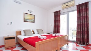 Ein Hotelzimmer mit einem hölzernen Bett, einem rot-weiß karierten Boden, einem Gemälde an der Wand und einem Balkon mit gemusterten Vorhängen.