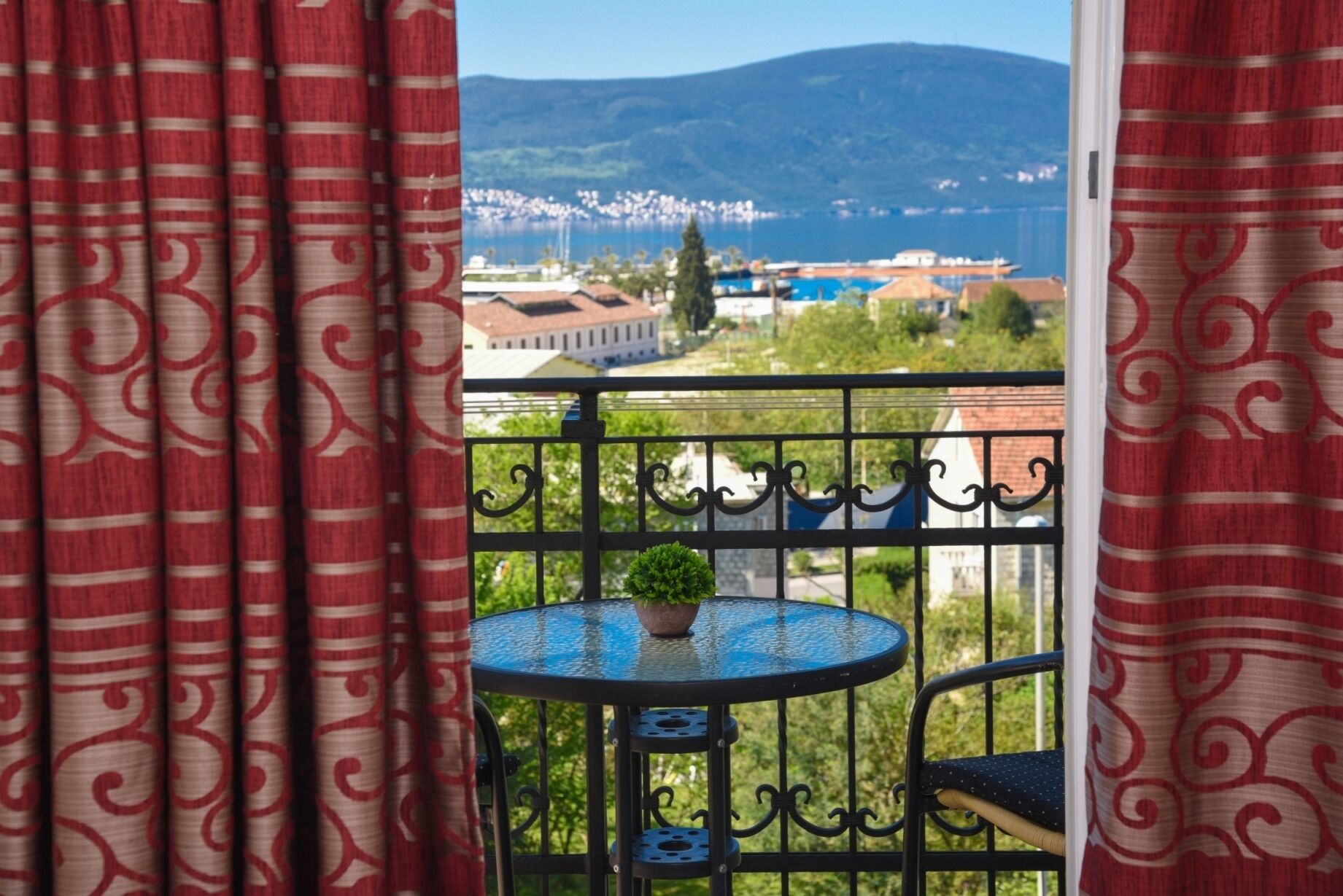 A balcony with a round table and a potted plant, overlooking a coastal town with buildings and a mountain in the distance.