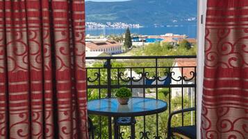A balcony with a round table and a potted plant, overlooking a coastal town with buildings and a mountain in the distance.