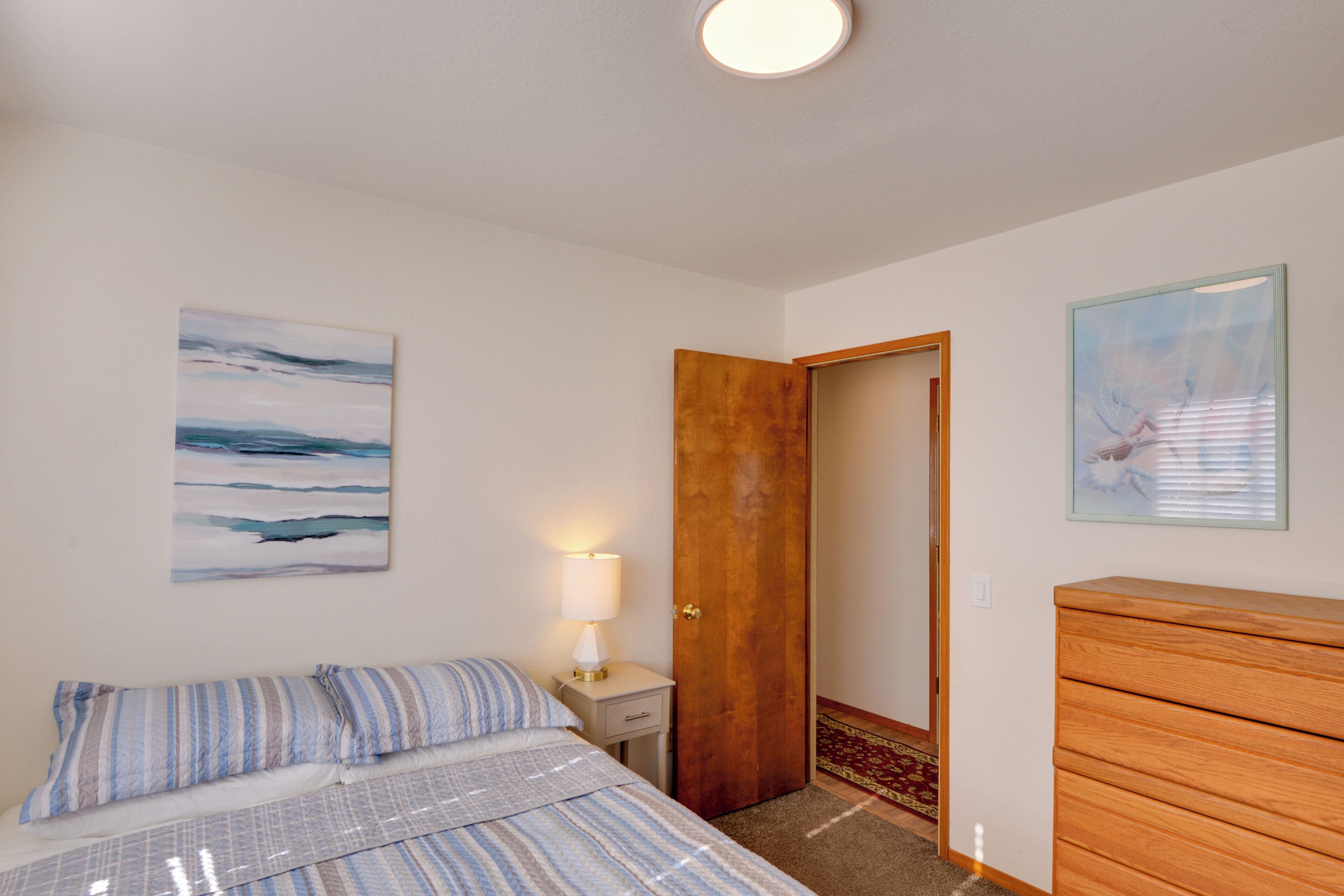 Une chambre à coucher avec un lit, une table de chevet avec une lampe, une commode en bois et deux tableaux accrochés aux murs.
