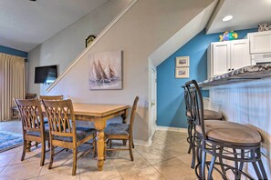 A dining area with a wooden table and chairs, a TV mounted on the wall, and a staircase leading to another room. - Steps to Sand! Miramar Unit w/ Pool, Tennis & More (Miramar Beach)
