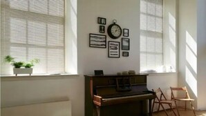A room with a piano, framed pictures on the wall, and a window with blinds. - Central Mezzanine Apartment in Historic School (Edinburgh)